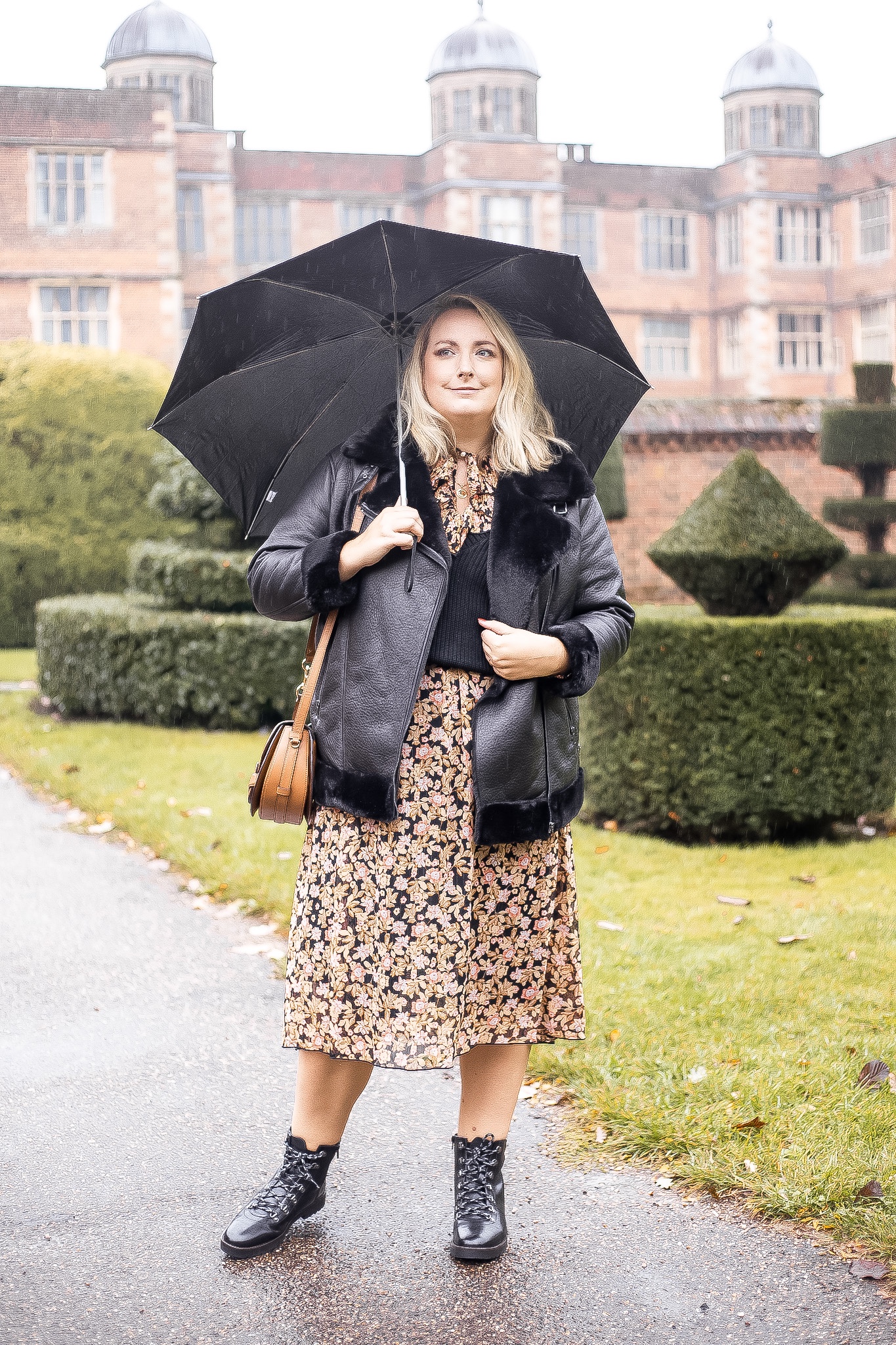 Lucy is stood in front of doddington hall, smiling holding an umbrella. She’s wearing a black jacket, dress and sweater vest
