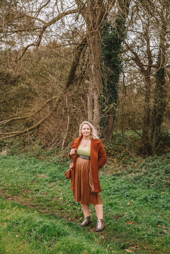 Lucy stood in a field with trees behind and wearing a pleather skirt, lime green knitted top and boots