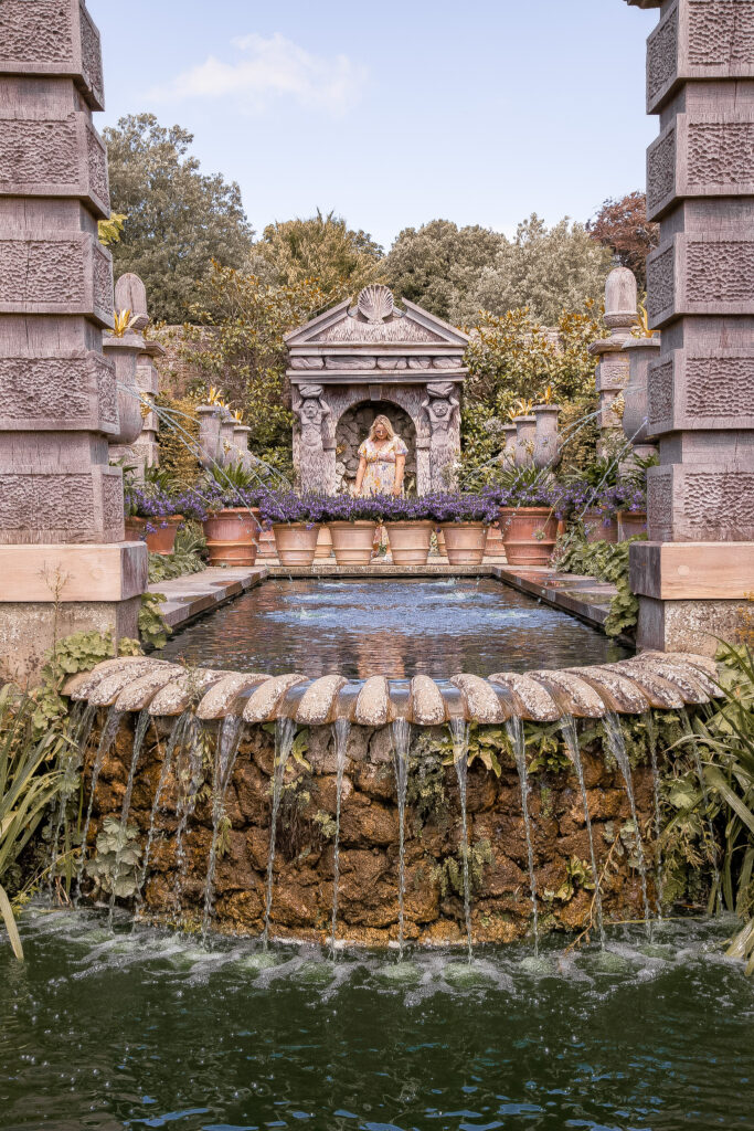 a large stone pond, has vases pouring water from their centres into it in an arch, and then this pours in streams into a smaller base pond. lucy is stood behind lavendar filled pots at the back