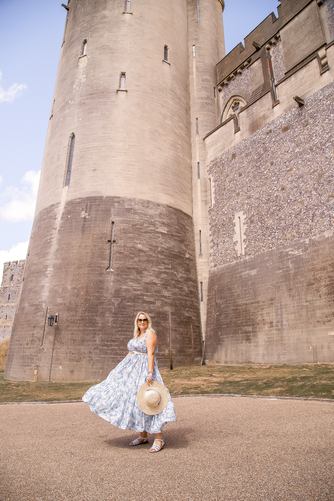 lucy is swisshing her maxi dress and holding a straw hat in front of a massive tower section of the castle