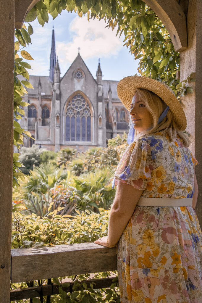 lucy is wearing a straw hat and pastel maxi dress and elaning on a wooden arch way with the church in the back ground framed by the wood and foliage