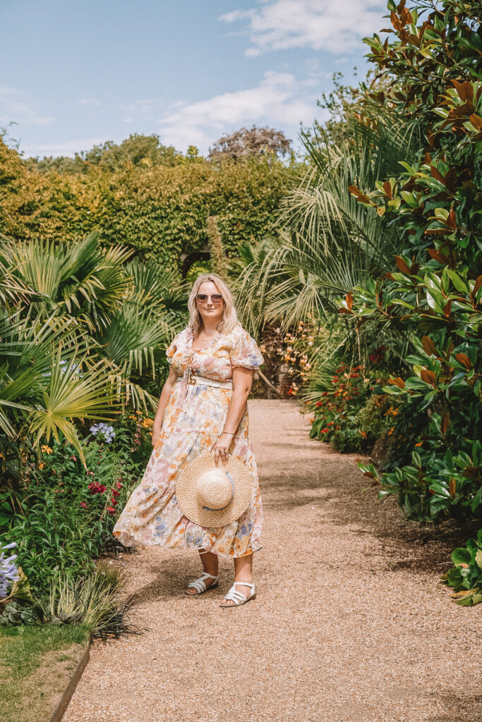 lucy in a pastel maxi dress holding a straw hat is stood on a path with exotic plam trees and other plants around her