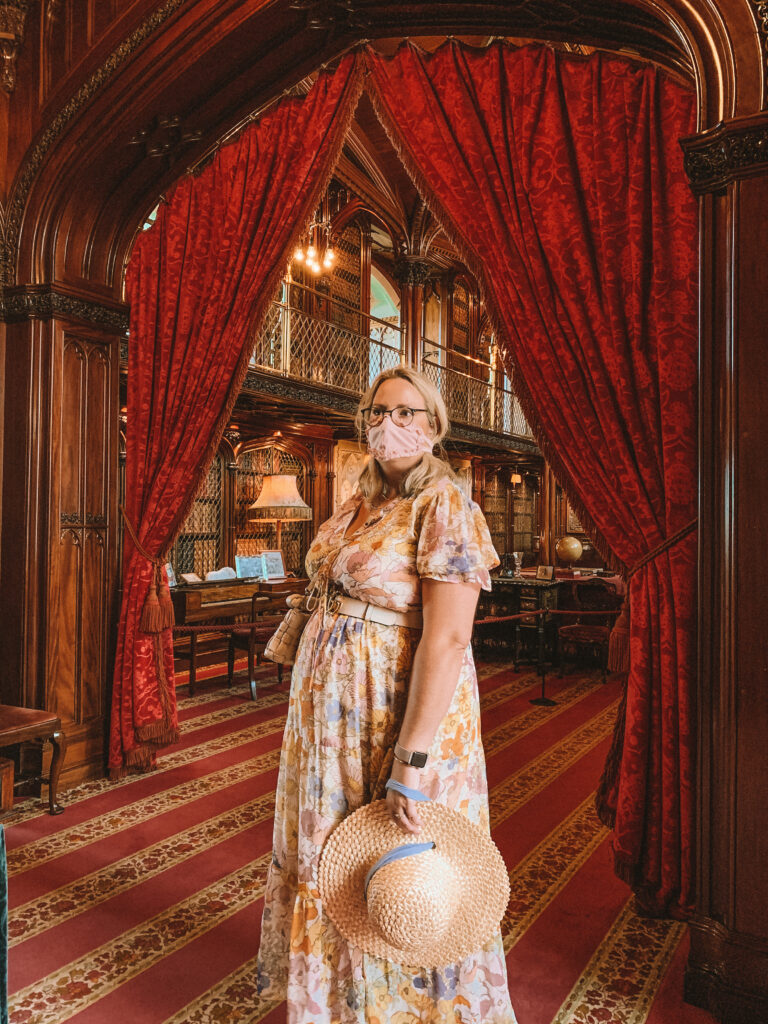 lucy is stood in a pastel maxi dress clutching a straw hat and wearing a face mask and glasses. shes stood in the library room with books and cabinets behind and a pair of big red curtains behind