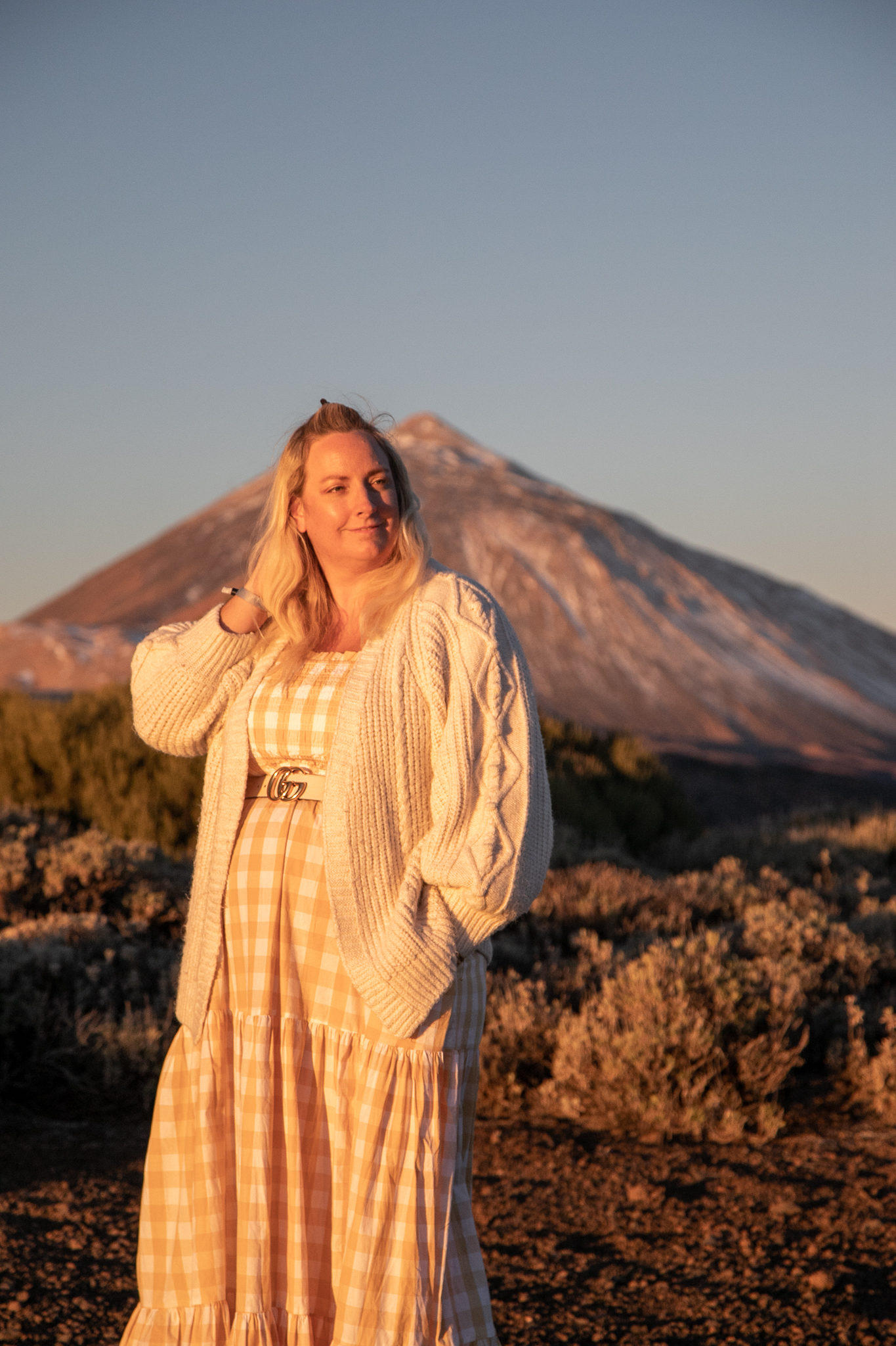 Lucy is stood with a yellow gingham dress and cream chunky cardigan on. The sun is shining on her and mount teide is in the background with snow on it